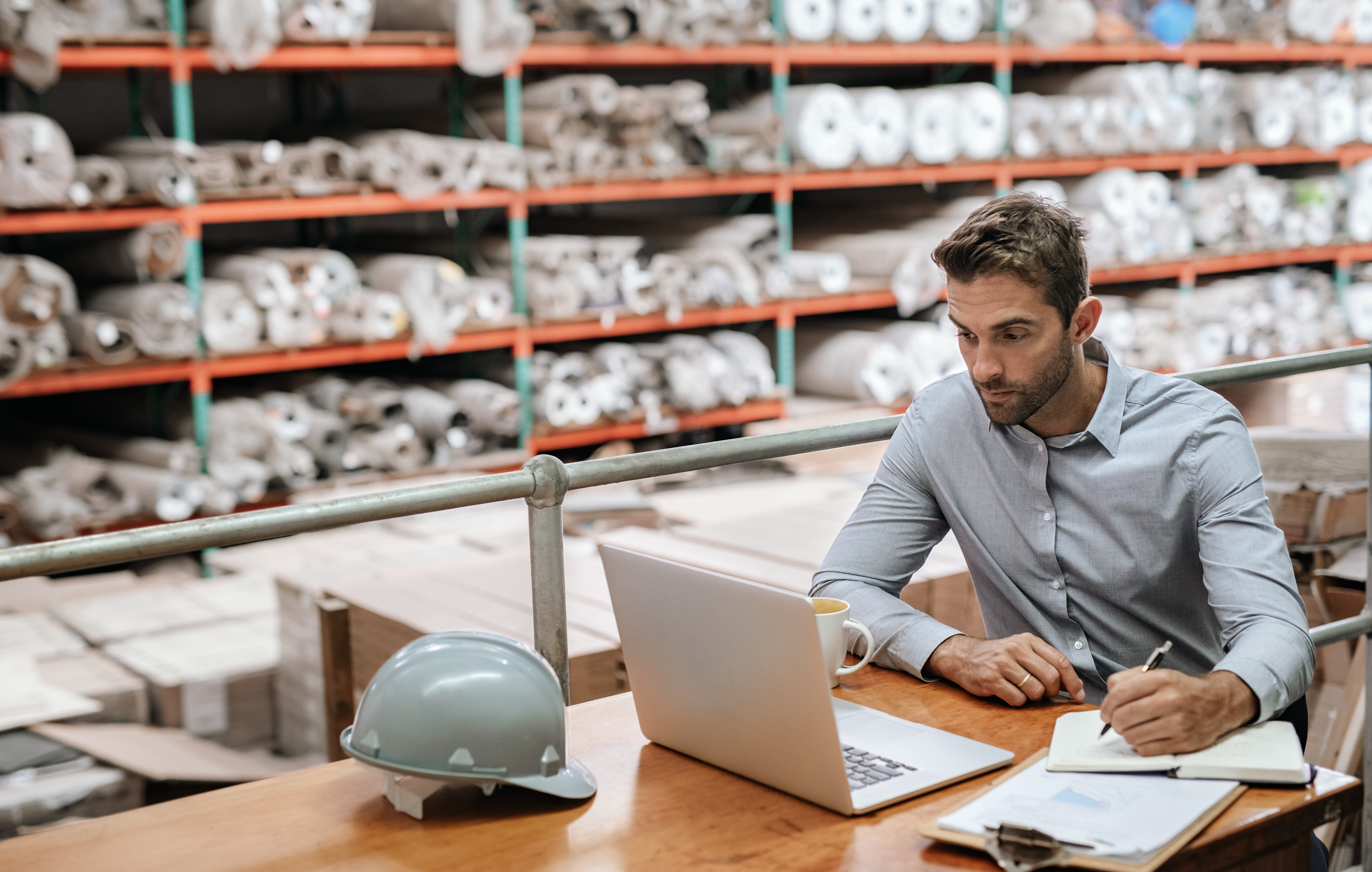 businessman sitting in a warehouse office looking at laptop and writing notes